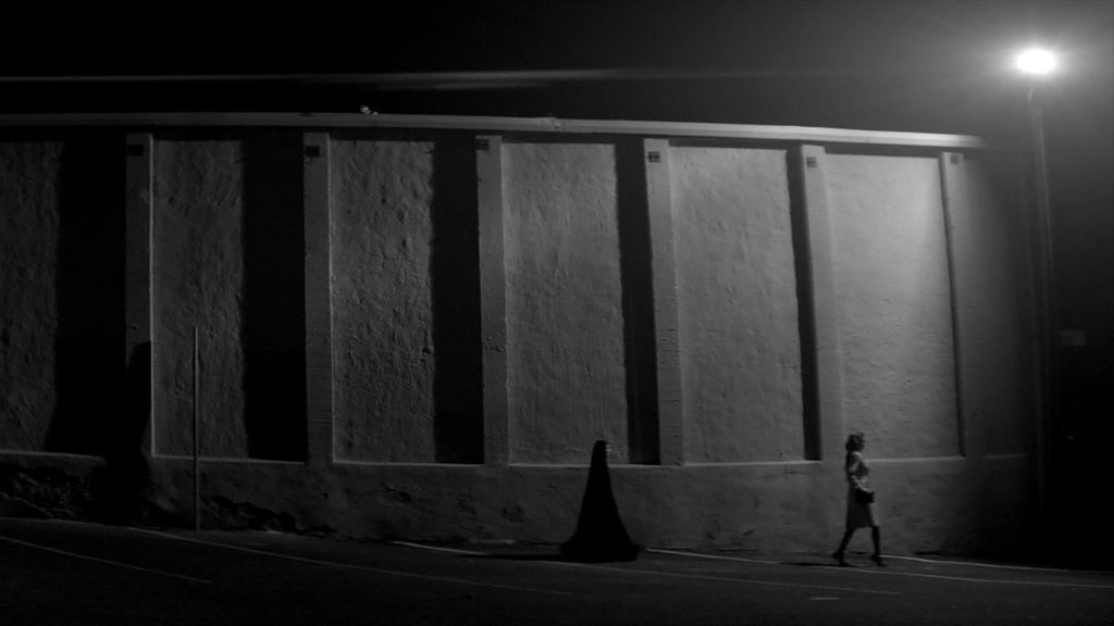 A wide shot of The Girl following a woman down the street.