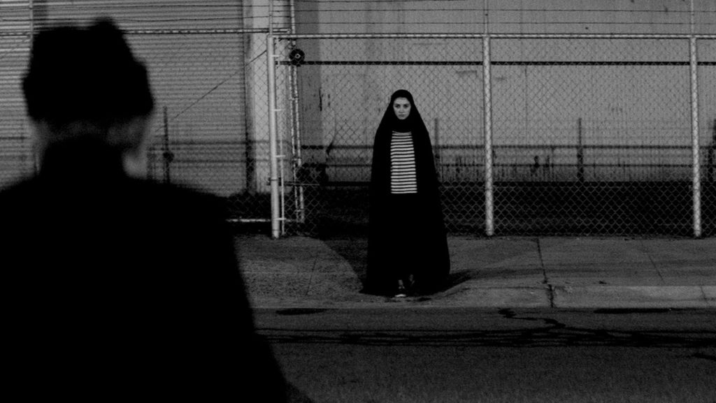 The Girl (Sheila Vand) watches a man from across the street. She is a young woman wearing a chador.