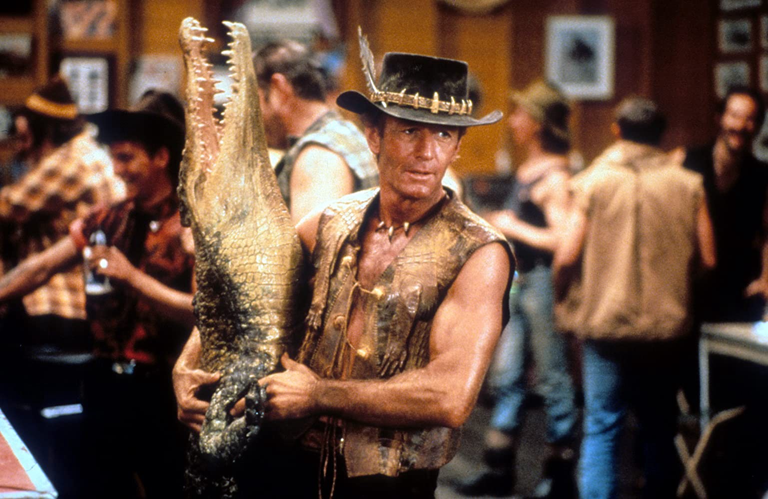 Mick, wearing an open leather vest and a feathered hat, holds a taxidermy crocodile