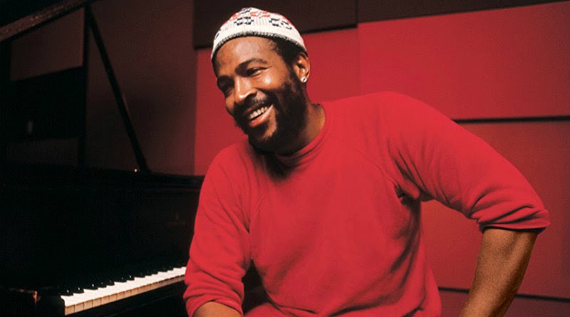 Marvin Gaye sits in front of a piano. He wears a beanie, a red sweater, and a bright smile.