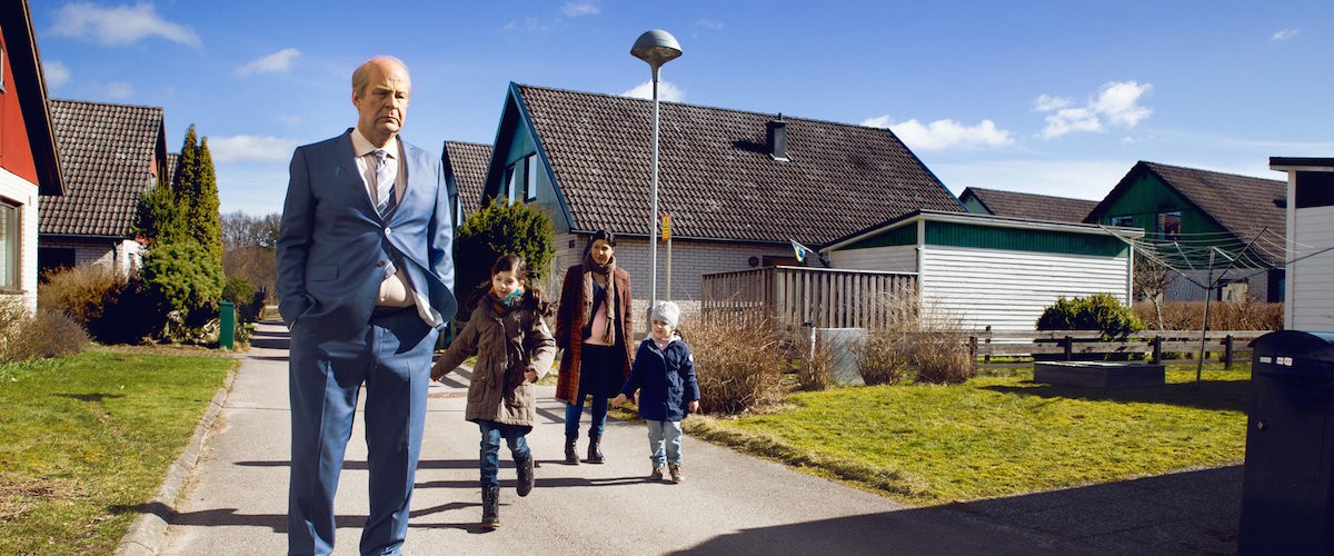 A man in a suit stands in front of a suburban home. A woman and two children are behind him, with one child running toward him with a smile on her face.