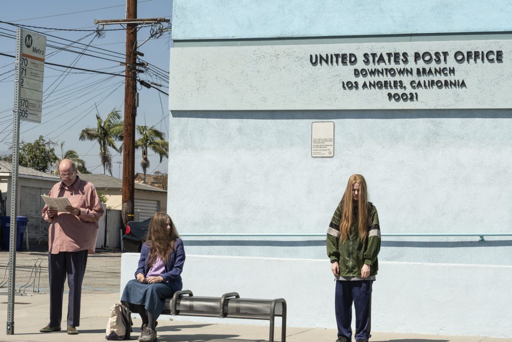 Robert, Theresa, and Old Dolio wait at a bus stop. Old Dolio stands apart from her parents and appears upset.