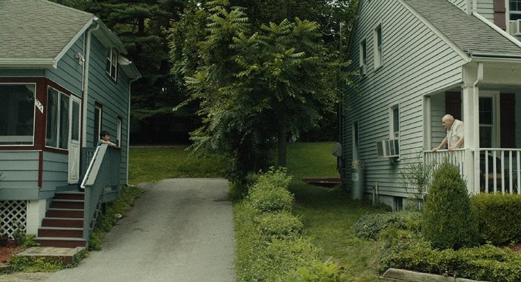 Two pale blue houses stand across from each, divided by a driveway and a lush green law. Del stands on one of the house's porch.