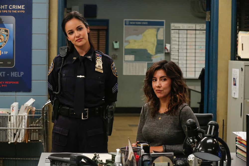 On the left, Melissa Fumero as Amy. She's standing and wearing a police uniform. Rosa, played by Stephanie Beatriz, sits to her right wearing civilian clothing.