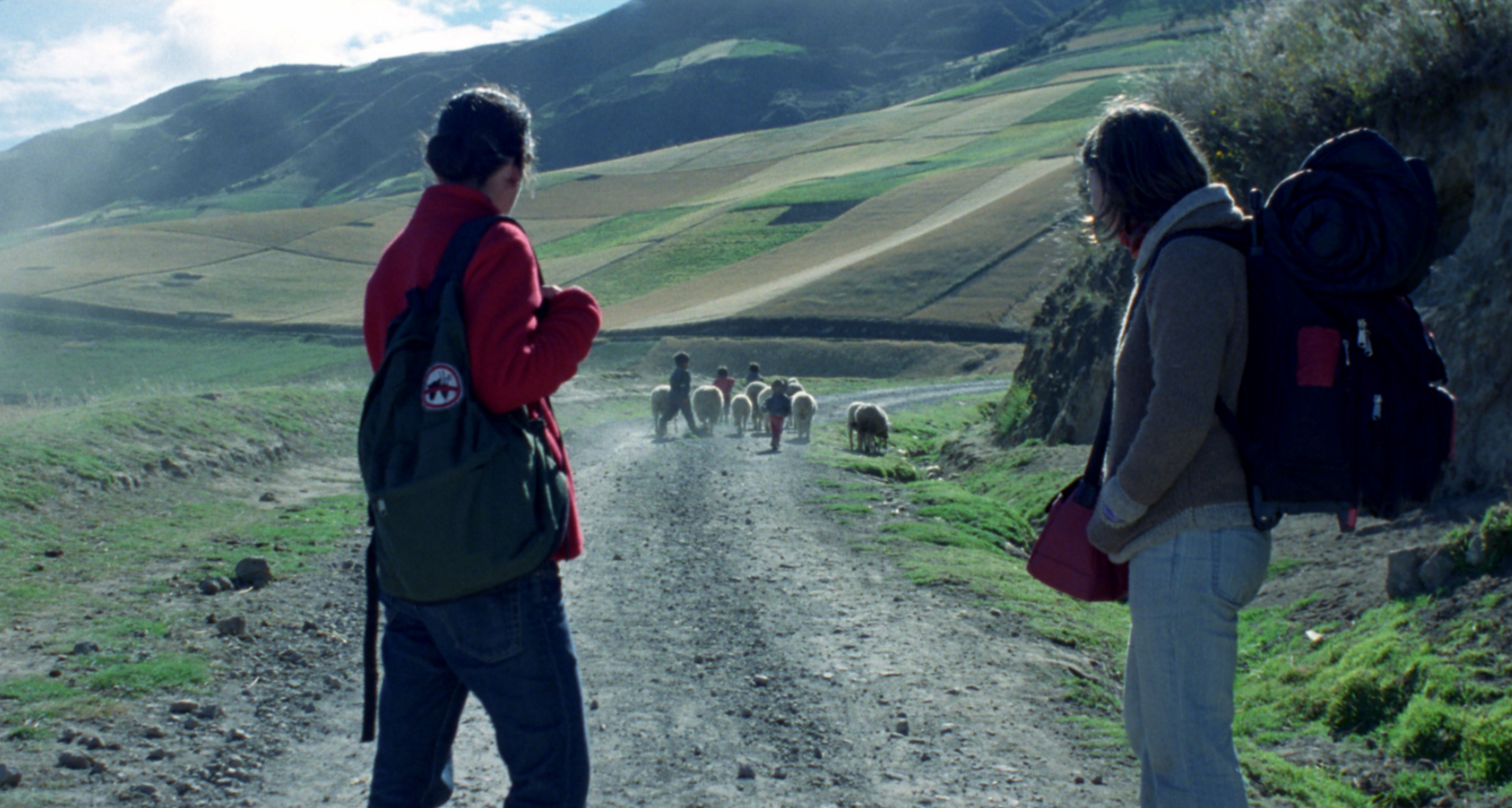 Two hikers observe a small herd of sheep and several children walking down a dusty road