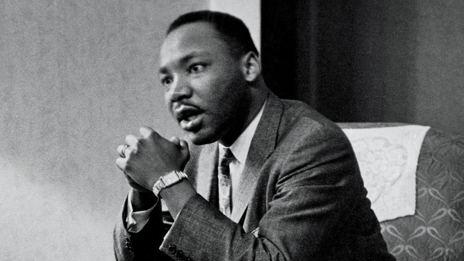 A black and white photo of Dr. Martin Luther King Jr. He is seated with his hands clasped in front of him, talking to someone to the left of the frame.