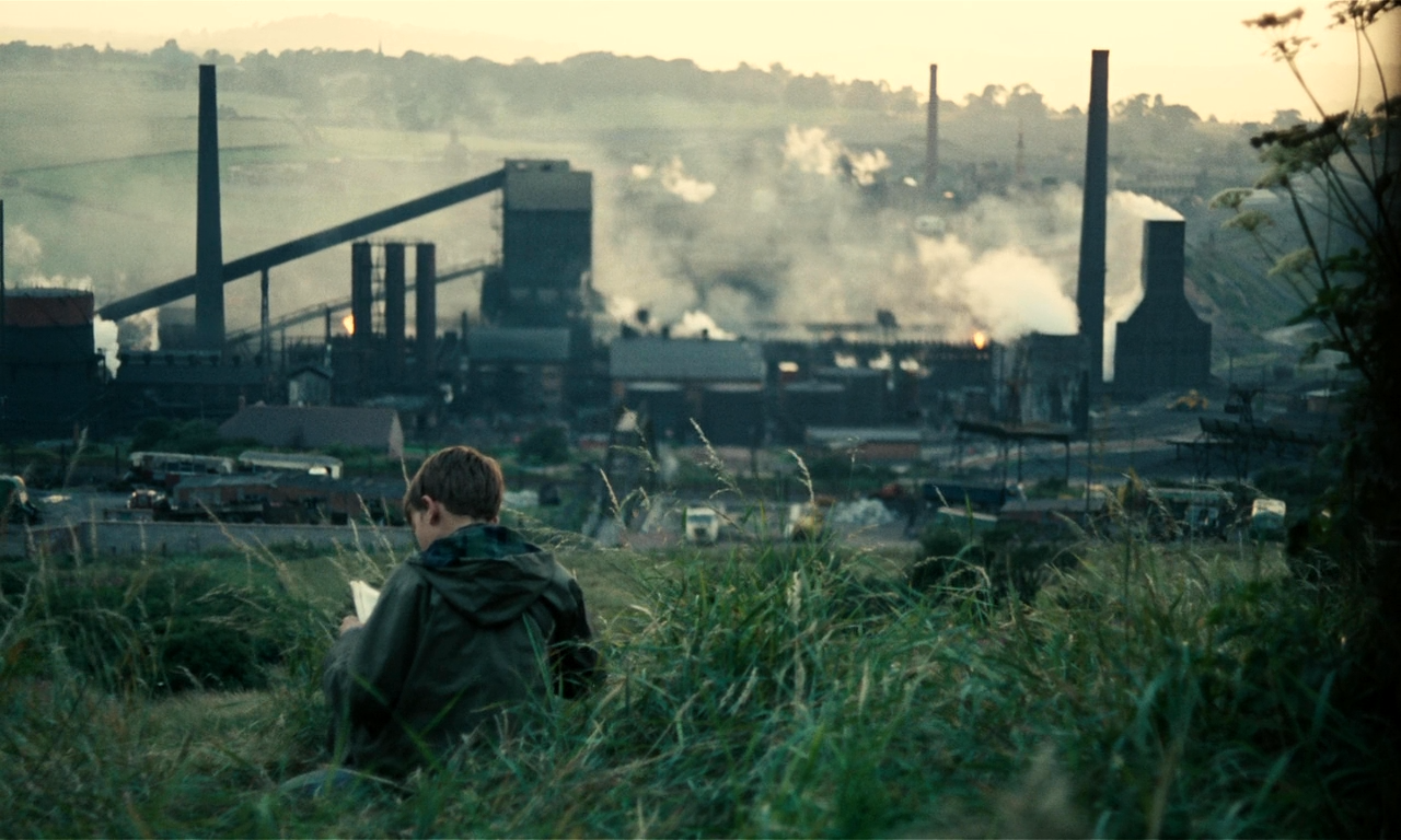 A young boy in a raincoat sits in a grassy field overlooking a factory