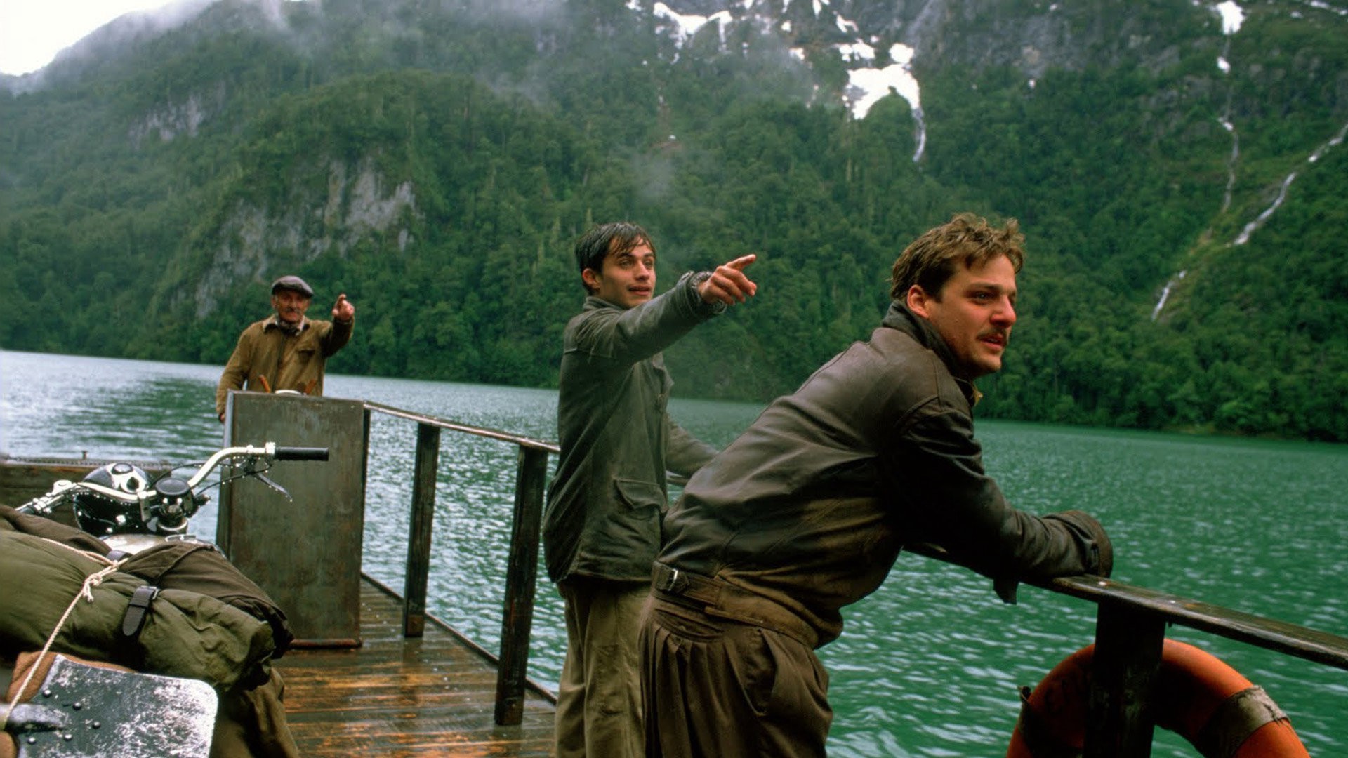 Three men ride down the river on a ferry. Two of the men are pointing to the right of the frame.