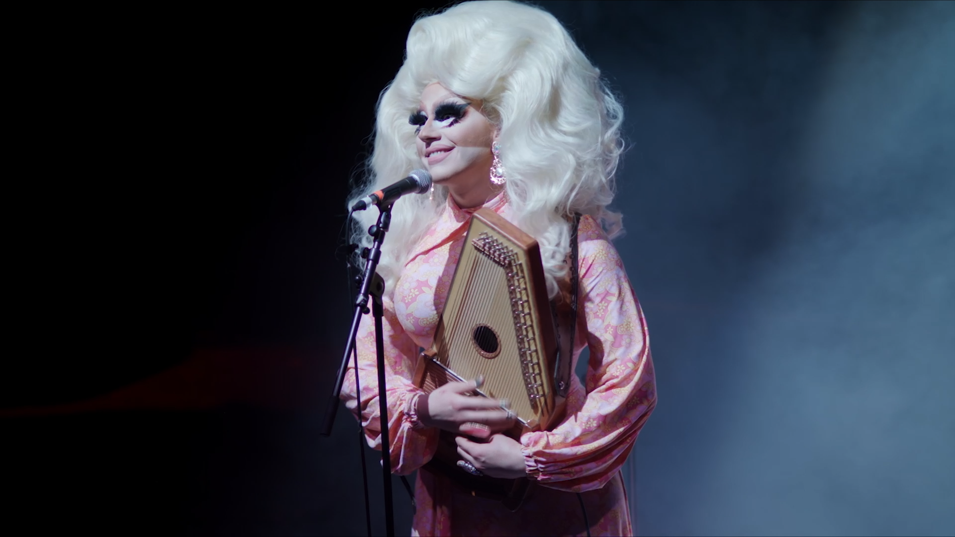 Trixie sings into a microphone and plays an autoharp. She wears a huge platinum blonde wig and a pink floral dress.
