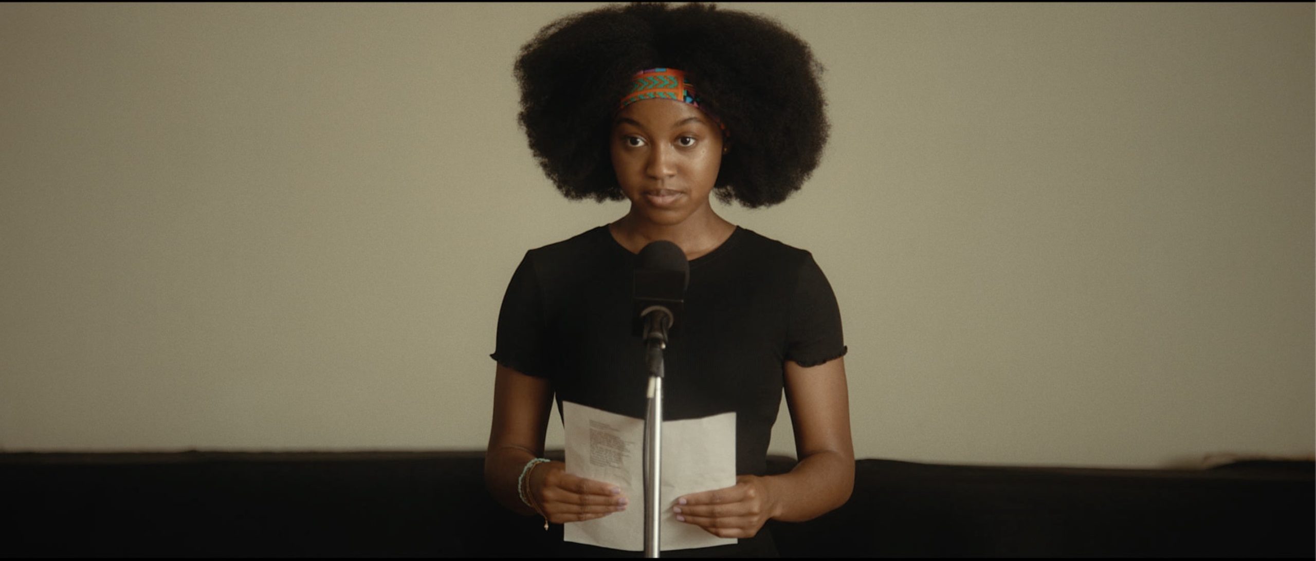 Kai stands in front of a microphone, holding a sheet of paper. She wears a black T-shirt and a headband, and wears her hair as an afro.