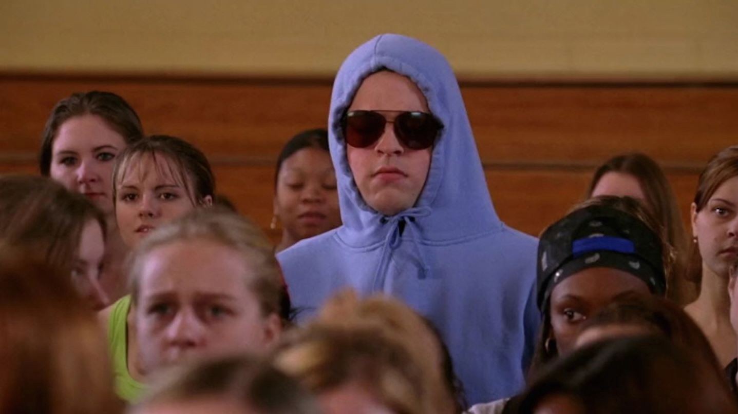 Damian from Mean Girls,  standing among students in a gym, wearing a lilac hoodie and sunglasses.