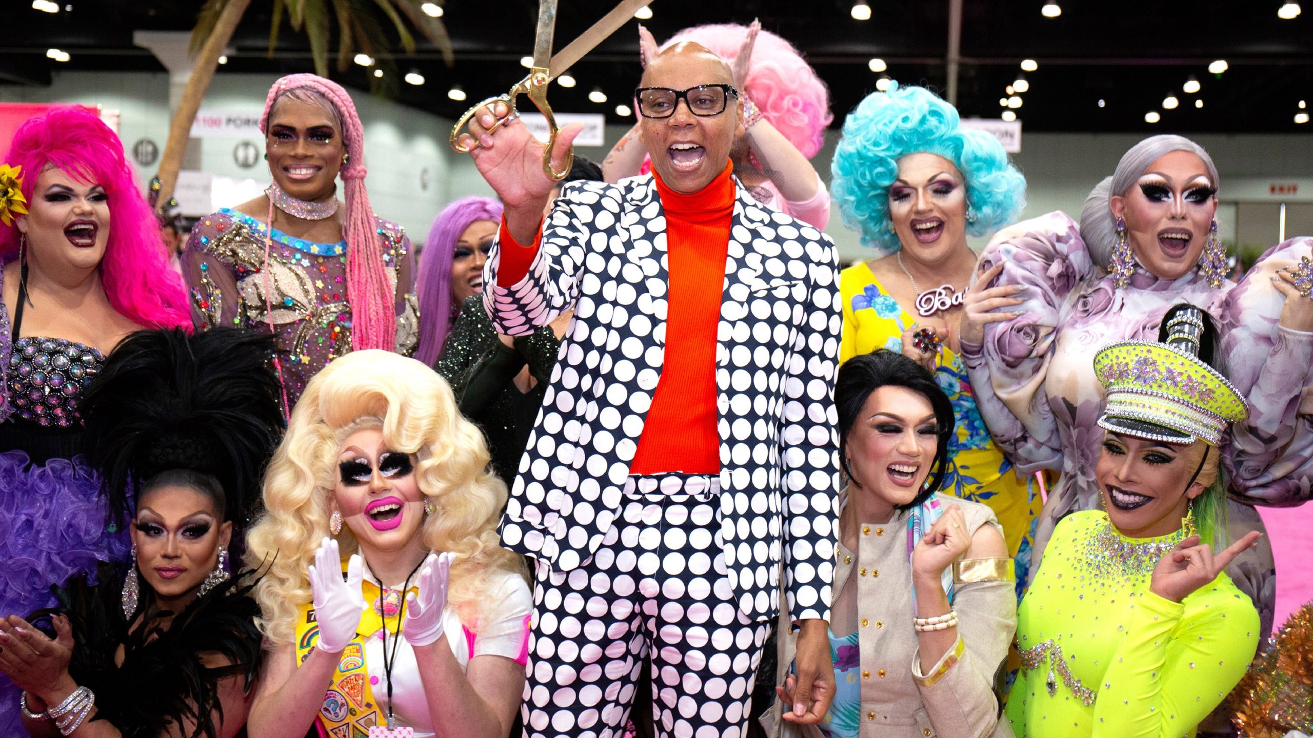 Rupaul, out of drag in a checkered suit, holding a pair of a golden scissors. A crowd of drag queens cheer behind her, including Trixie.