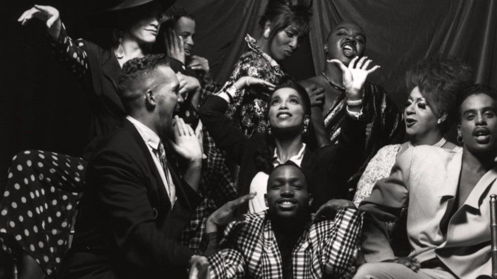 A black and white photo of a group of drag and ball performers. Some pose for the camera, and others laugh and smile at each other.