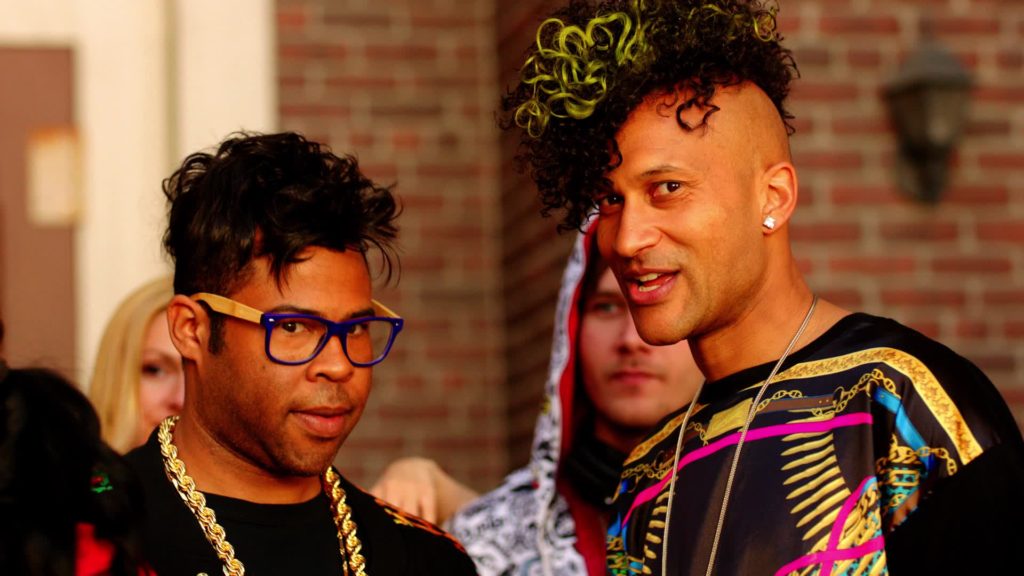 A still from Key & Peele's "Nooice" sketch, with the two standing shoulder-to-shoulder in a small crowd. Peele wears blue and yellow hipster glasses and a thick gold chain and has an uncomfortable expression on his face. Key has a curly yellow-streaked mohawk, wears a brightly colored shirt, and is smiling.