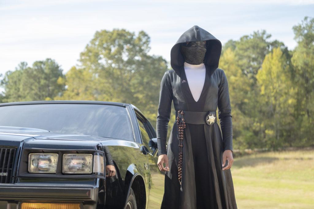 Regina King as Angela Abar in Watchmen. She stands in front of a black car, wearing a long black cloak with a sheriff's badge on the waist and a black mask that entirely covers her face.