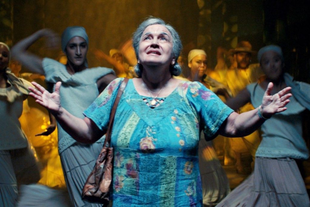 Abuela Cluadio, an older woman with grey hair and a blue dress, stands in front of a group of female dancers. Her arms are open and she looks to to the sky.