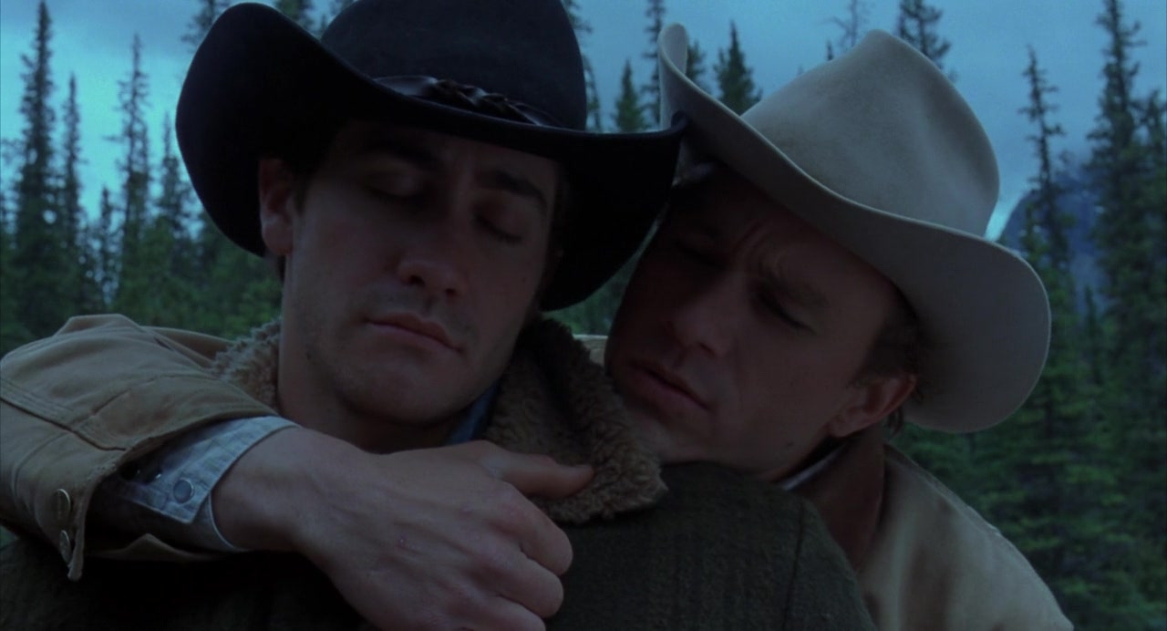 Ennis (Heath Ledger) embraces Jack (Jake Gyllenhal) from behind. Both wear cowboy hats and have their eyes closed. A forest is behind them.