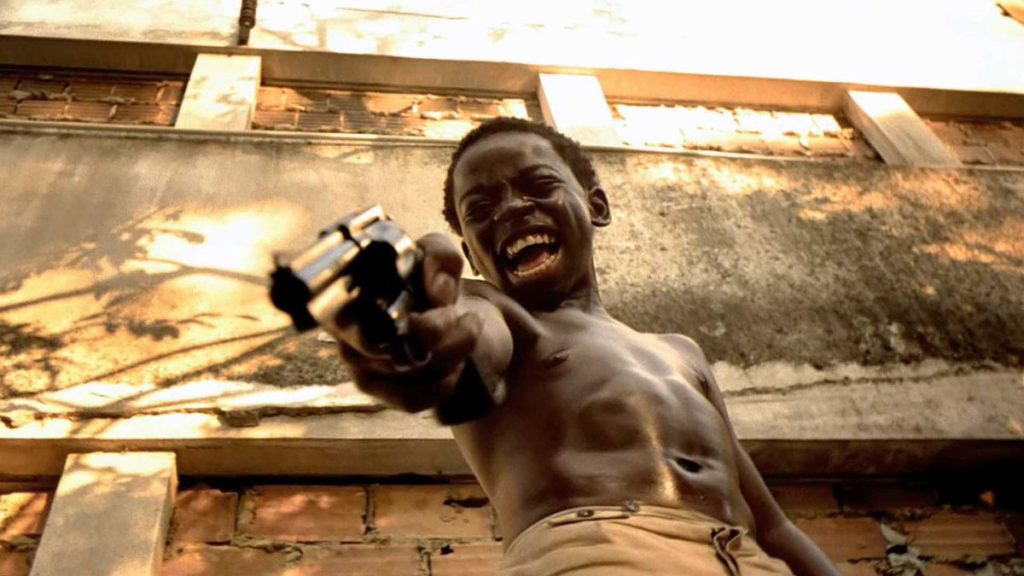Douglas Silva as Li'l Dice. He's a young boy, shirtless and wielding a gun against a concrete backdrop.