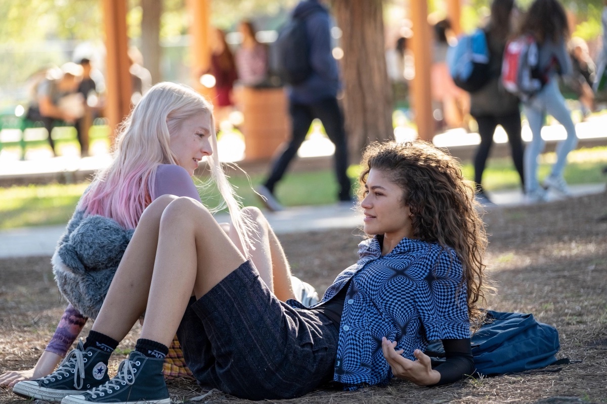 Jules (Hunter Schafer) and Rue (Zendaya) relax and chat in a park.