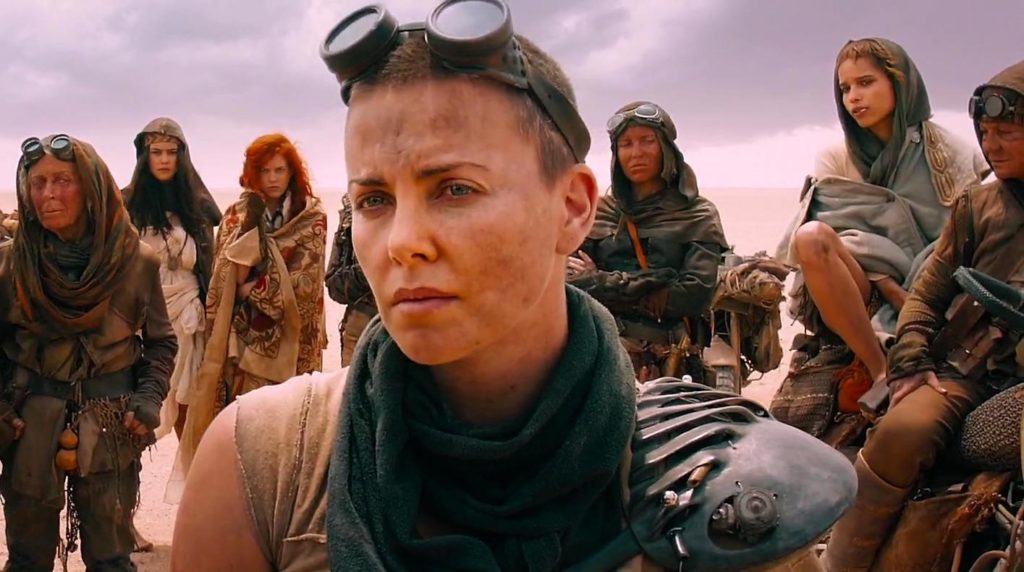 Furiosa (Charlize Theron) stands in the foreground with a pair of goggles on her forehead. A group of women stand behind her. In the background, a vast desert and a cloudy sky.