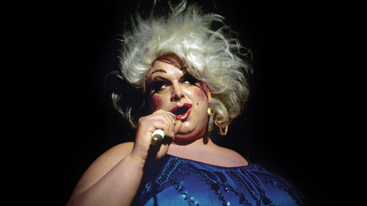 Divine, a drag queen wearing exaggerated makeup and a blue dress, speaks into a microphone.