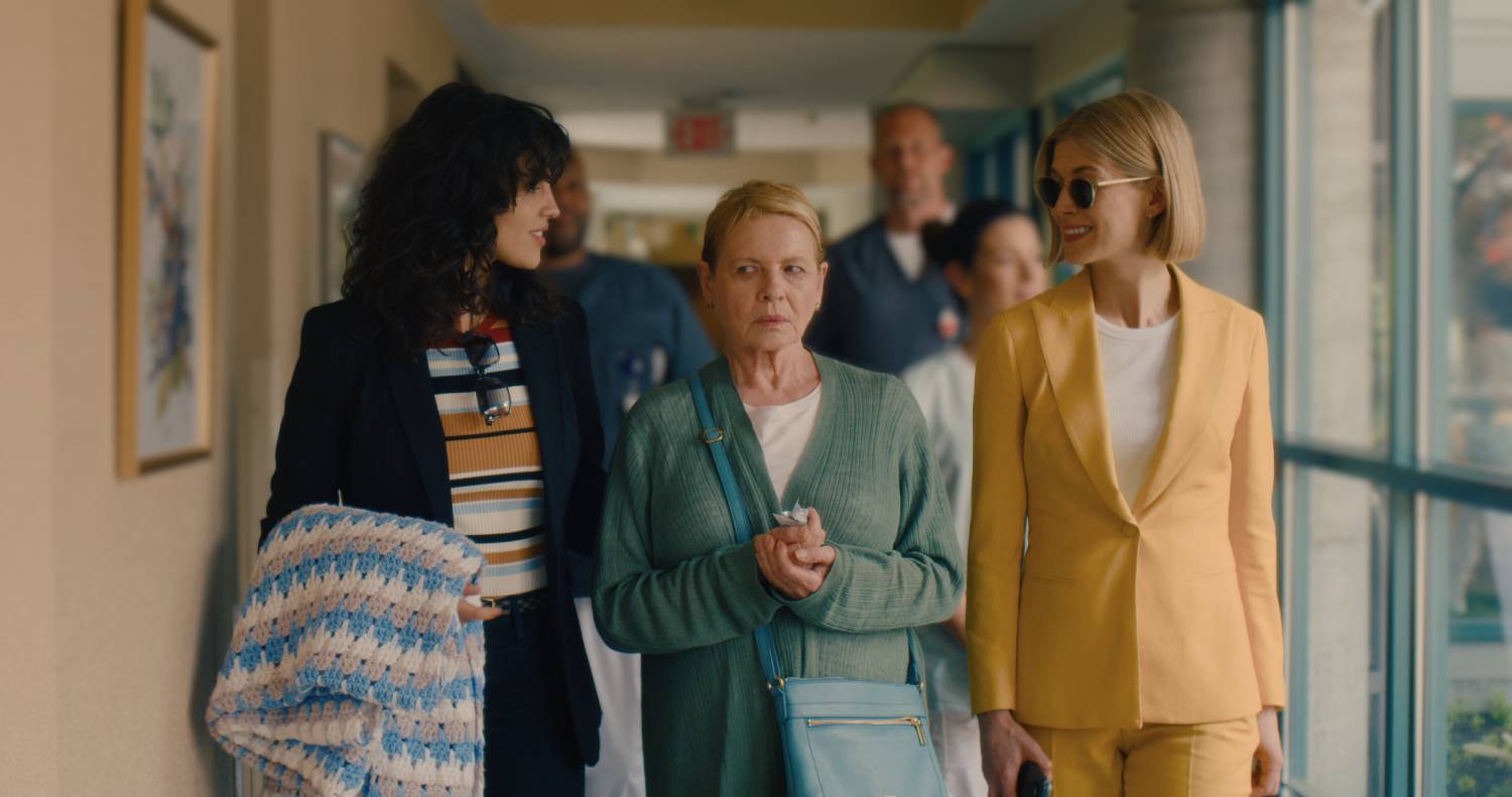 Marla and Fran (two young women) walk Jennifer (an older woman) down a hallway. Marla and Fran are smiling, and Jennifer looks suspicious.