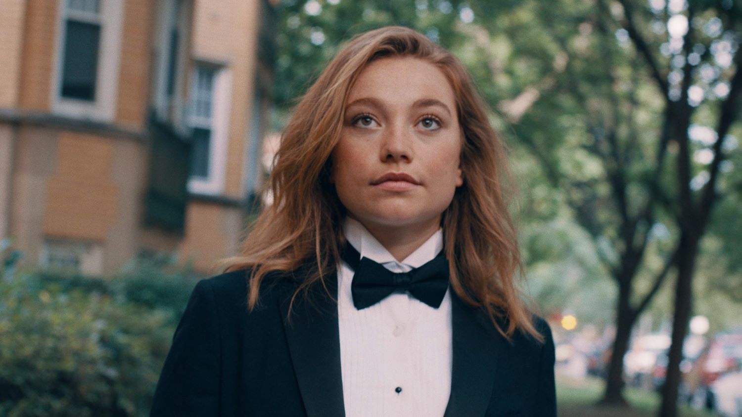 Cyd stands on a city block in front of a tree. She's a young woman with long brown hair wearing a tuxedo. 