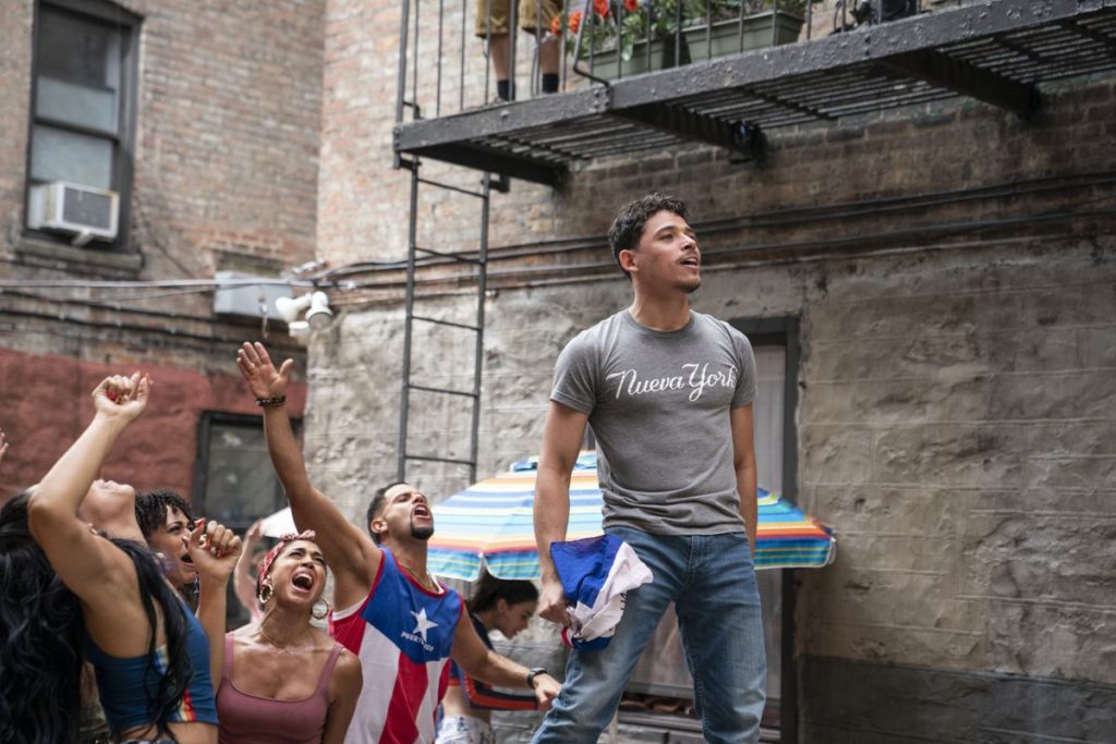 Usanavi stands above a cheering crowd. He wears a T-shirt that reads "Nuevo York" and holds a crumpled flag in one hand.