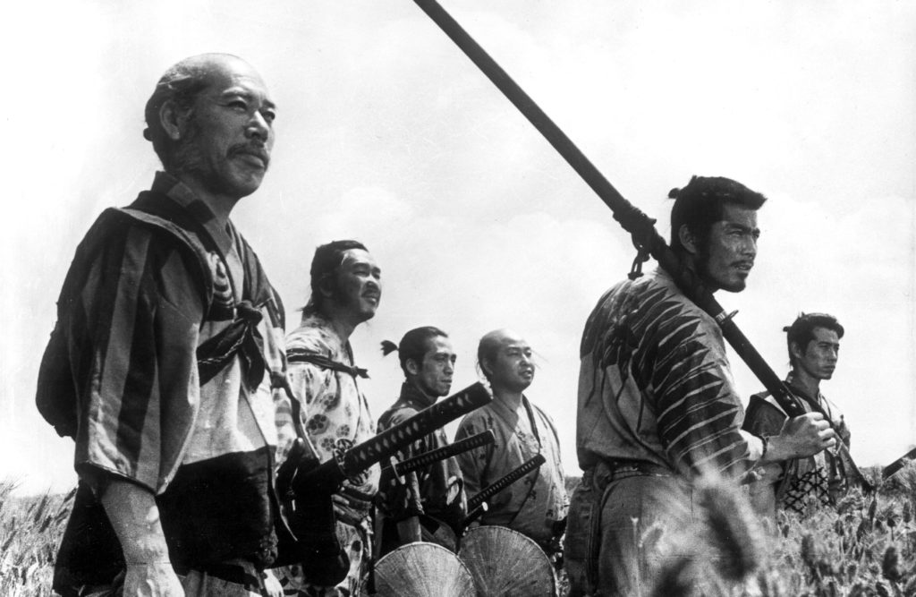 The seven samurai stand in a field. Kambei (Takashi Shimura) and Kikuchiyo (Toshiro Mifune) are in the foreground of the image. Kambei has a slight smile on his face, and Kikuchiyo holds an enormous katana over his shoulder.