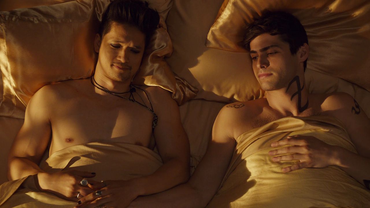 Magnus and Alec lie in bed, shirtless, holding hands over the sheets.