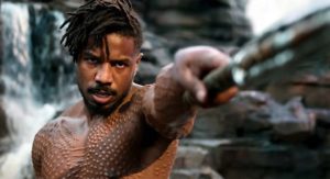 A Black man scowls and points a spear at the camera. He is shirtless, revealing many scars.