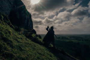 Large shot of the sky and the mountain side. Gawain is walking downward with an axe in his hand. 