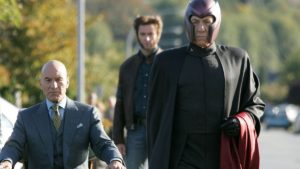 A bald man in a wheelchair and an old man in a cape and helmet walk towards the camera, followed by a man with spiky hair.