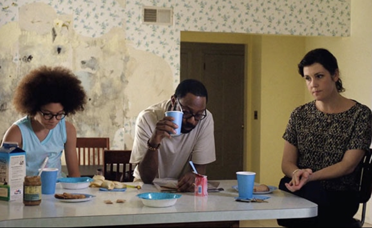The Burns family sitting at the kitchen table for breakfast. There is friction between them all, shown by their expressions. Black mold is exposed beneath the wallpaper behind them.