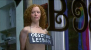 A still from Better than Chocolate of Maggie protesting naked with a sign that says "obscene lesbian" over her breasts
