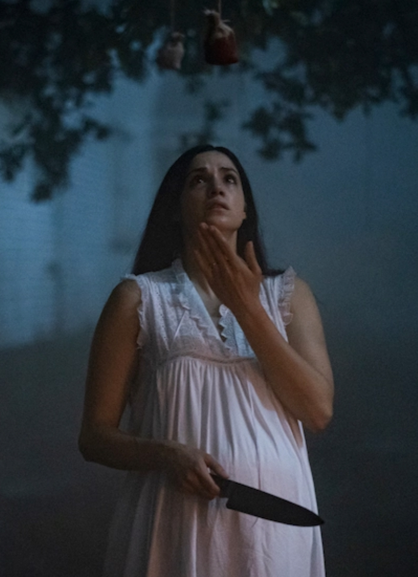 A woman in a white nightgown with long dark hair is pictured outside at night. She holds a large knife, looking upwards at bloody pouches hanging from a tree.