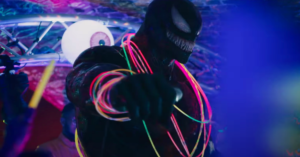 A still from "Venom: Let There Be Carnage" of Venom at a rave, wearing glow bands around his neck and arms, dropping a mic