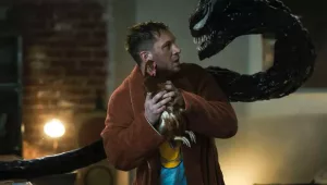 A still from "Venom: Let There Be Carnage" of Eddie, looking fearful and holding a chicken, and Venom's head popping out of his back and looking at him