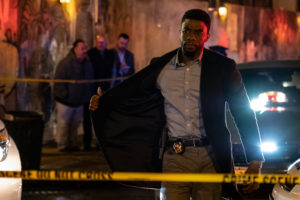 Chadwick Boseman in "21 Bridges". He holds his jacket open to show his badge, and is surrounded by do not cross tape.