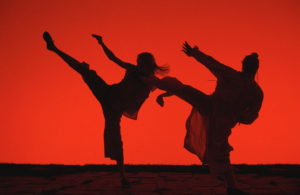 Two people standing in a fighting pose, one leg and arm in the air, against a red background.