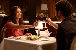 A still from 'Love Hard' of Natalie and Tag at a dinner table in a fancy restaurant, clinking wine glasses
