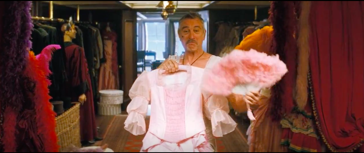 A still from Stardust of Robert De Niro as Captain Shakespeare smiling in a dress, holding a pink dress and pink fan in front of him