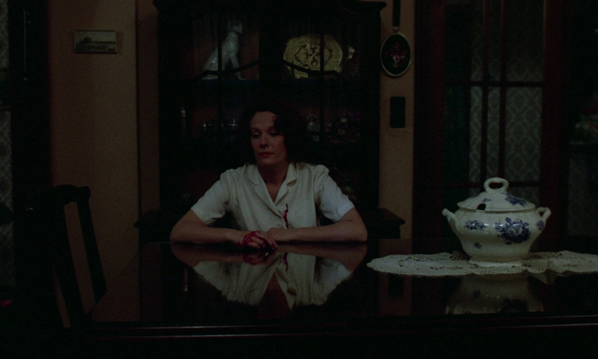 Jeanne Dielman in a dim-lit room, she has blood covered on her hands and shirt. She's sitting at table, soup tureen is present