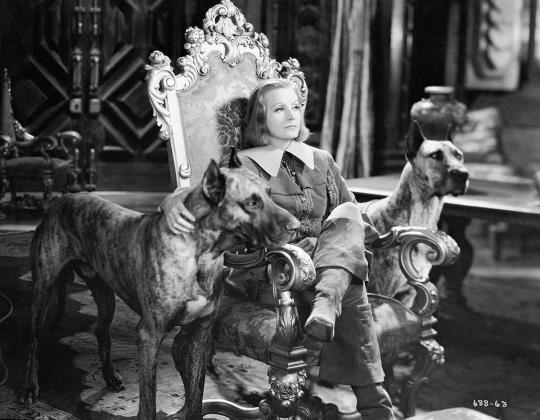 Christina sitting on a throne chair, wearing men's clothes near two big dogs