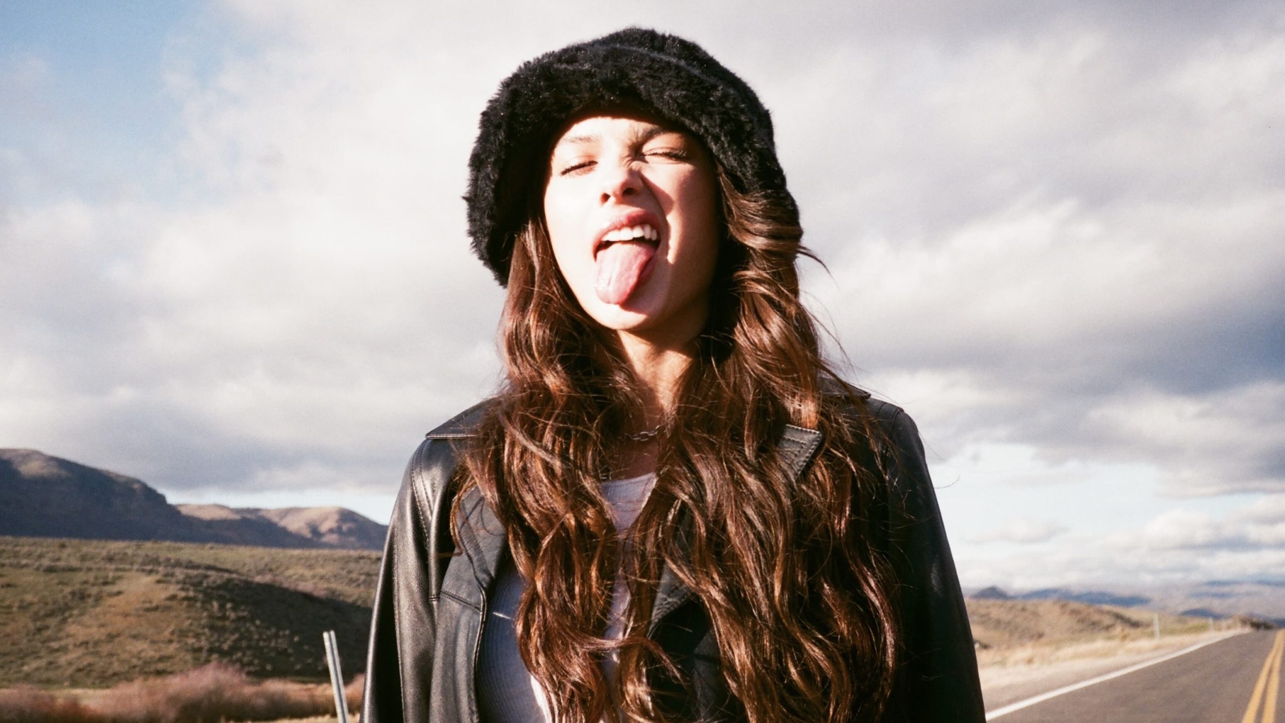 olivia rodrigo wearing a black jacket, she's sitcking her tongue out in a pose