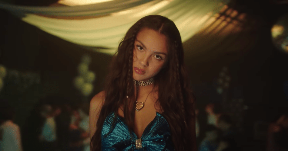 Close-up of Olivia Rodrigo wearing a blue dress, in a populated school gym