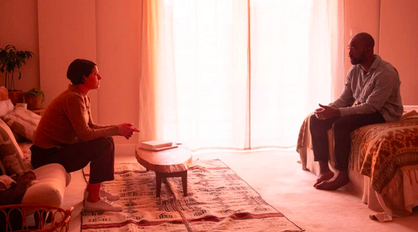 A woman and man sit across the room from each other. The frame is very warm, almost orange in tone.