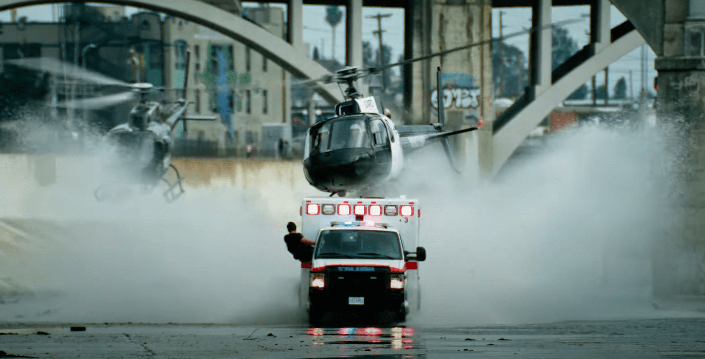 Ambulance rushing through water, helicopter closing in on top, man sticking out from ambulance