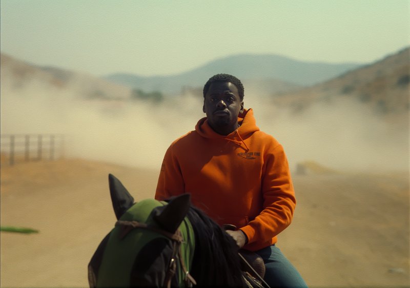 An image of Daniel Kaluuya as OJ in an orange hoodie and snapback on a horse in a dusty setting