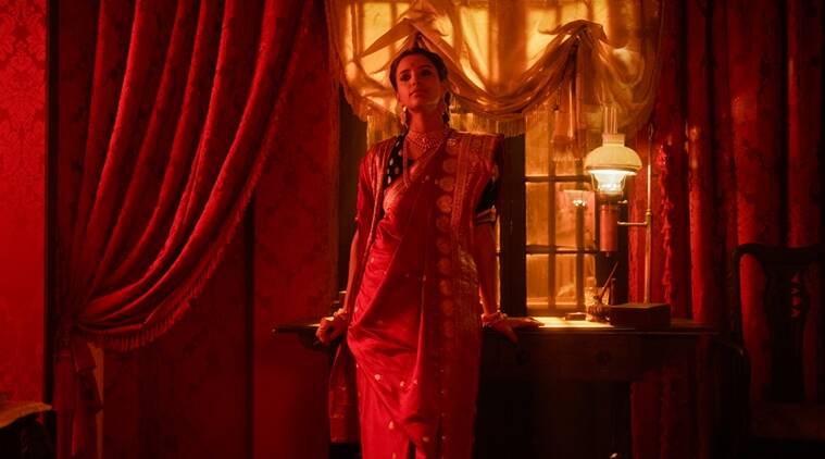 Bulbbul is wearing a red indian sari dress. She's standing near a small table, near red drapes